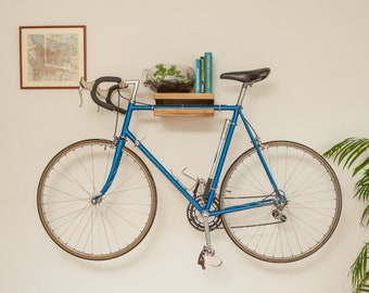 May include: A blue bicycle is mounted on a wall with a wooden shelf above it. The shelf holds a small glass terrarium with green plants and a few books. The bicycle has a black seat and black handlebars.