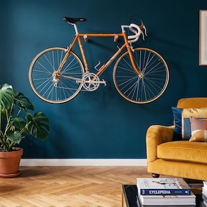 May include: An orange road bicycle mounted on a dark teal wall above a mustard yellow armchair. The room features a monstera plant in a terracotta pot, a small side table, and framed line art prints hanging on the wall above the chair.