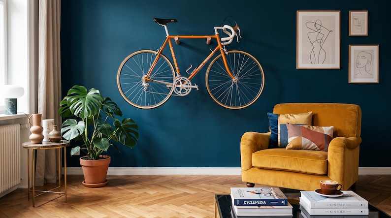 May include: An orange road bicycle mounted on a dark teal wall above a mustard yellow armchair. The room features a monstera plant in a terracotta pot, a small side table, and framed line art prints hanging on the wall above the chair.