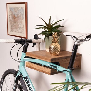 May include: A light blue Bianchi Nirone 7 road bike mounted on a rectangular wooden wall shelf. The shelf holds a gold pineapple ornament and a green potted plant. A framed map hangs on the wall above the bike, which features white handlebar tape.