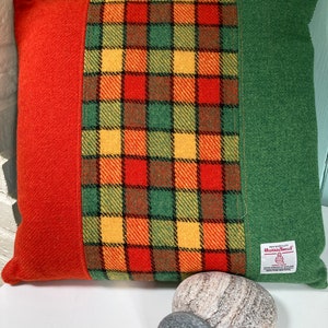 Puede incluir: Una almohada de color rojo, verde y amarillo a cuadros con un borde verde y una etiqueta "Harris Tweed". La almohada está sobre una superficie blanca con dos piedras grises.