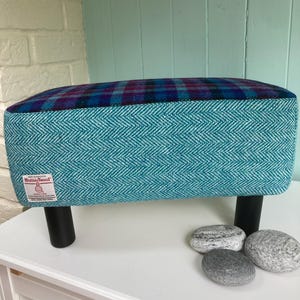 May include: A rectangular footstool with a teal herringbone fabric on the sides and a plaid top in blue, purple and teal. It has black legs and a Harris Tweed label. The dimensions are approximately 40 cm x 30 cm.