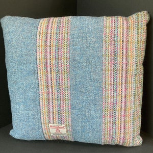 May include: Square blue tweed pillow with a central vertical stripe of colorful woven fabric. The pillow has a tag that reads "Harris Tweed". The pillow is a decorative home accent.