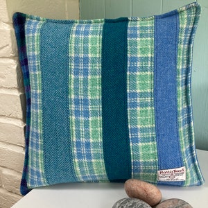 May include: A blue, green, and white plaid throw pillow with a "Harris Tweed" label. The pillow is sitting on a white surface with three gray stones in front of it.
