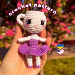 Könnte beinhalten: Handgemachte Häkelpuppe in Form einer Teetasse mit lila Tutu. Die Puppe hat einen weißen Kopf mit schwarzen Augen und rosigen Wangen. Der Text "crochet pattern" ist über der Puppe gebogen.