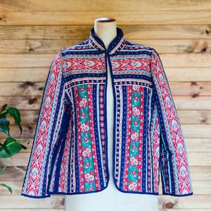 May include: A quilted jacket with a vibrant floral and geometric pattern in red, blue, and white. The jacket features a mandarin collar and a vertical stripe design with floral accents. The jacket is displayed on a mannequin against a wooden background.