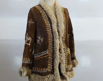 May include: A brown suede coat featuring a thick beige shearling trim along the collar, cuffs, and hem. The coat is decorated with white and gold embroidered patterns, including wavy lines and star-like motifs on the front and sleeves.