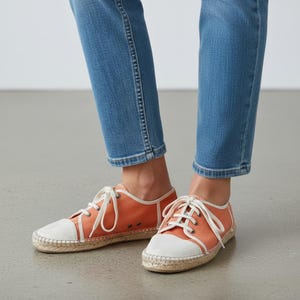 Puede incluir: Zapatos espadrilles de lona naranja con punteras blancas y cordones blancos. Los zapatos tienen una suela de yute tejida y ribete blanco. Llevados con vaqueros azules.