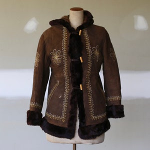 May include: A brown suede coat with a hood and faux fur trim. The coat features floral embroidery and wooden toggle closures. The cuffs and hem are trimmed with dark brown faux fur, creating a vintage aesthetic.