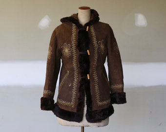 May include: A brown suede coat with a hood and faux fur trim. The coat features floral embroidery and wooden toggle closures. The cuffs and hem are trimmed with dark brown faux fur, creating a vintage aesthetic.