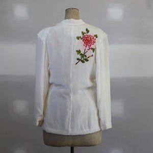 May include: Cream-coloured long-sleeved blouse with a floral embroidery on the back. The embroidery features a large red and pink flower with green leaves and stems. The blouse is displayed on a mannequin.