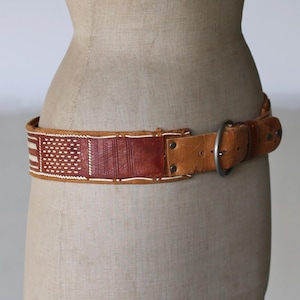 Puede incluir: Un cinturón de cuero marrón con un panel decorativo tejido con detalles en blanco y rojo. El cinturón tiene una hebilla grande de color plateado y se muestra en un maniquí. El cinturón es de estilo ancho.