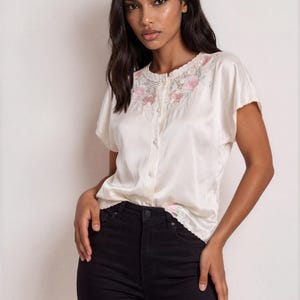 Puede incluir: Una blusa de manga corta color crema con botones, que presenta un delicado cuello de encaje con detalles florales bordados en rosa. La blusa tiene un acabado suave tipo satén y se combina con unos vaqueros negros de tiro alto.