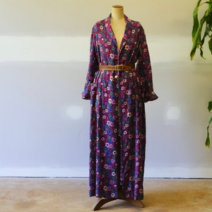 Puede incluir: Una larga bata de estampado floral morado con un cinturón marrón. La bata tiene cuello en V y mangas largas con volantes en los puños.