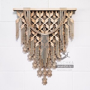 Puede incluir: Un tapiz de pared de madera con un patrón geométrico y flecos. El tapiz está hecho de madera marrón claro y tiene un acabado natural. El tapiz mide aproximadamente 61 cm de ancho y 91 cm de alto. El tapiz está firmado "Nischka makes" y "Nischka.com".
