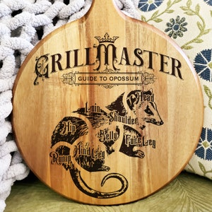 May include: Round wooden cutting board with a handle, featuring the text "GRILL MASTER GUIDE TO OPOSSUM" and an illustration of an opossum with labeled cuts of meat. The board is made of light brown wood.