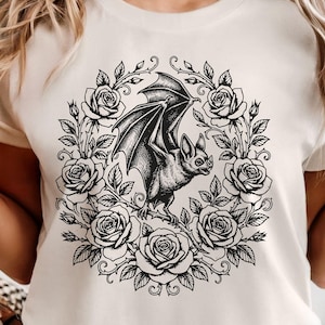 May include: Ivory-colored t-shirt featuring a black and white illustration of a bat within a floral wreath of roses. The word "IVORY" is printed at the bottom. The shirt has short sleeves and a crew neck.