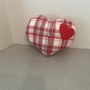 May include: Heart-shaped pillow with a red and white plaid pattern. A small red heart is sewn onto the front. The pillow has a button closure on the side. A decorative item for a bedroom or living room.