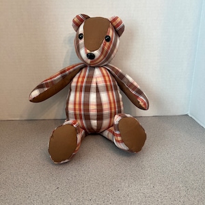 May include: A handmade teddy bear with a plaid pattern in shades of brown, red, and cream. The bear has brown paws, ears, and a snout. The bear is sitting upright, with its arms and legs extended.