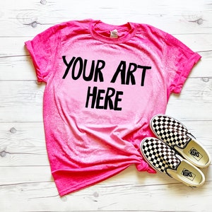 May include: A pink t-shirt with a faded ombre effect, featuring the text "YOUR ART HERE" in black. The shirt is laid flat on a white wooden surface, with a pair of black and white checkered Vans slip-on shoes beside it.