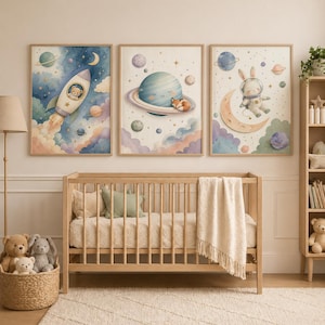 May include: Three framed watercolour art prints with a space theme. One shows a rocket with a bear, another a planet with a fox, and the third a bunny on a crescent moon. The prints are in a nursery setting with a cot and soft toys.