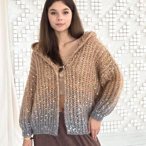 May include: A woman wearing a hooded cardigan with a gradient color scheme, transitioning from tan to gray. The cardigan is paired with brown corduroy pants. The background features a white wall with a geometric design and beige curtains.