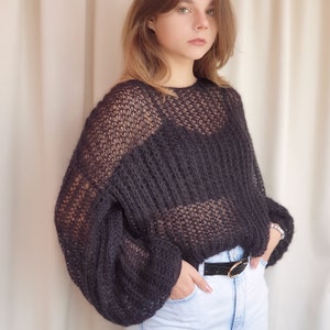 May include: A black, sheer, knitted sweater with a loose fit and long sleeves. The sweater has a delicate, open-weave pattern and a slightly cropped length.