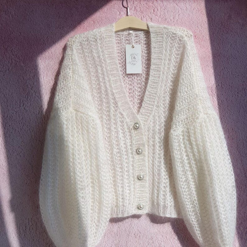 May include: White knitted cardigan with a V-neck and pearl buttons. The cardigan has a loose fit and long sleeves.