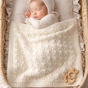 May include: A sleeping baby in a wicker bassinet, nestled on a pillow and covered with a cream-colored knitted blanket. The baby wears a matching bonnet and long-sleeved shirt. A wooden teether is placed on the blanket.