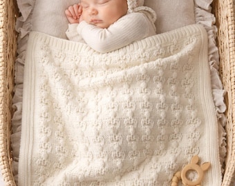 Könnte beinhalten: Ein schlafendes Baby in einer Weidenkorb-Wiege, auf einem Kissen und bedeckt mit einer cremefarbenen Strickdecke. Das Baby trägt eine passende Mütze und ein langärmliges Oberteil. Ein Holzbeißring liegt auf der Decke.