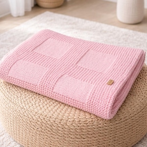 Può includere: Una coperta rosa chiaro lavorata a maglia con un motivo quadrato strutturato, ripiegata su un pouf beige intrecciato. È presente una piccola etichetta di legno. La coperta misura circa 127 cm per 152 cm.