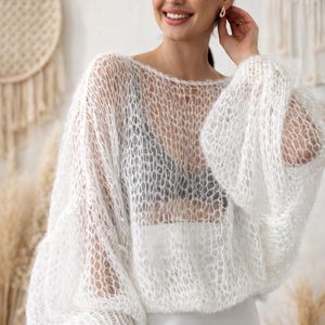 May include: A white, open-weave sweater with long, voluminous sleeves. The sweater has a round neckline and is paired with a white skirt. The style is airy and light, suitable for layering. The model is smiling.