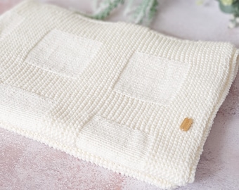 May include: A white knitted baby blanket with a square pattern. The blanket is folded and has a small wooden tag attached.