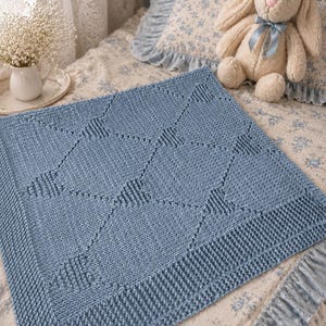 Könnte beinhalten: Hellblaue gestrickte Babydecke mit geometrischem Rautenmuster. Die Decke hat einen strukturierten Rand und wird auf einem Bett mit einem floral gemusterten Kissen und einem Stoffhasen präsentiert.