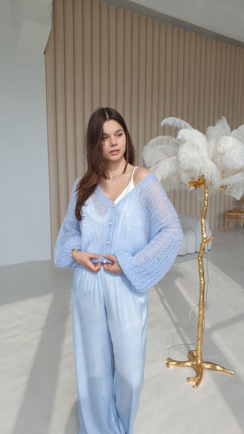 May include: A light blue knitted cardigan with pearl buttons and long sleeves. The cardigan is worn over a white tank top and light blue pants.