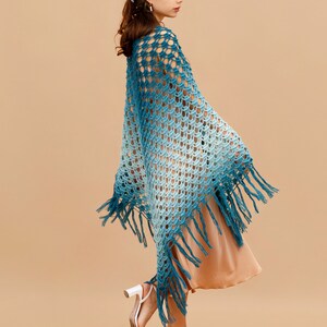 May include: A woman wearing a blue and white ombre crochet shawl with fringe. The shawl is a triangular shape and has a delicate openwork pattern. The woman is wearing a light brown dress and white heeled sandals.