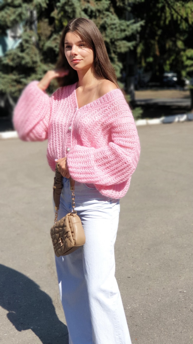 May include: A pink knitted cardigan with a button closure and long sleeves. The cardigan is worn over a pair of light blue jeans. The person is carrying a brown quilted handbag with a chain strap.