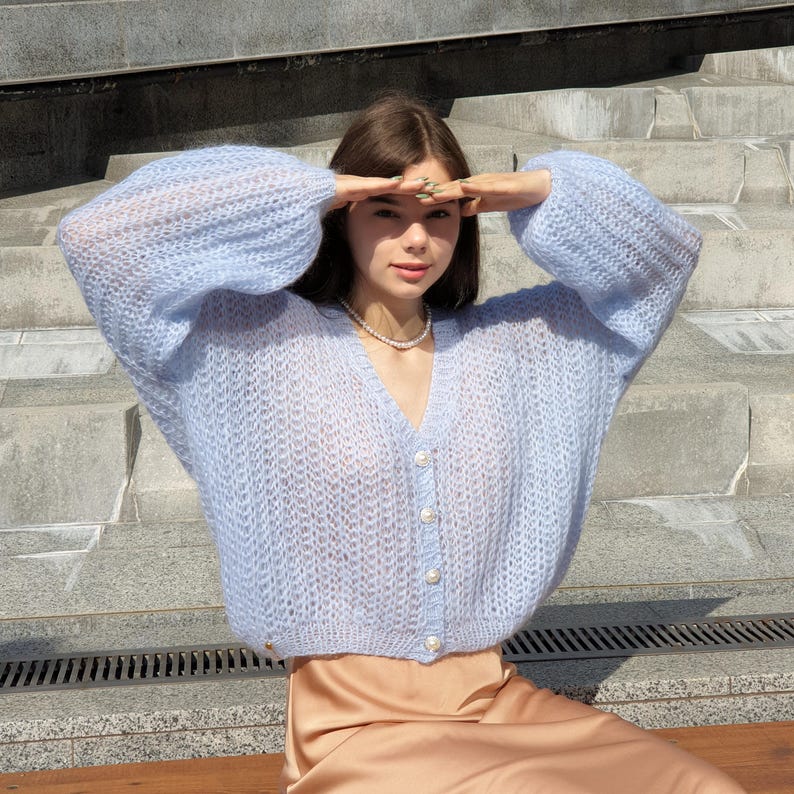 May include: A light blue knitted cardigan with pearl buttons and a V-neck. The cardigan is made of a soft, airy yarn and has a relaxed fit.