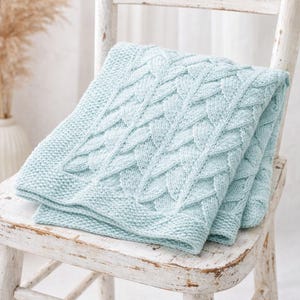 May include: A folded, light blue knitted blanket with a cable knit design rests on a distressed white wooden chair. The blanket features a textured pattern and a smooth border. The chair has a rustic, aged appearance.