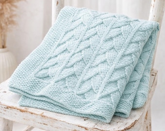 May include: A folded, light blue knitted blanket with a cable knit design rests on a distressed white wooden chair. The blanket features a textured pattern and a smooth border. The chair has a rustic, aged appearance.
