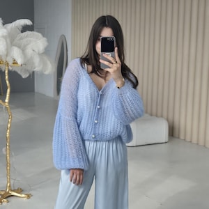 May include: A light blue knitted cardigan with pearl buttons and long, puffed sleeves. The cardigan is paired with matching light blue wide-leg pants.