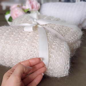 May include: A white crocheted baby blanket with a white satin ribbon tied in a bow. The blanket is soft and fluffy, perfect for a newborn.