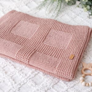 Può includere: Una morbida coperta per neonati in maglia rosa con un design strutturato. La coperta presenta un motivo quadrato al centro e un bordo a coste. È attaccata una piccola etichetta di legno. Un giocattolo di legno per bambini è nell'angolo.