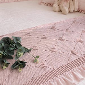 May include: A pale pink knitted blanket with a geometric design and a frilled edge. The blanket is on a bed with a matching pillow and a stuffed bunny. Green foliage is placed on the blanket.