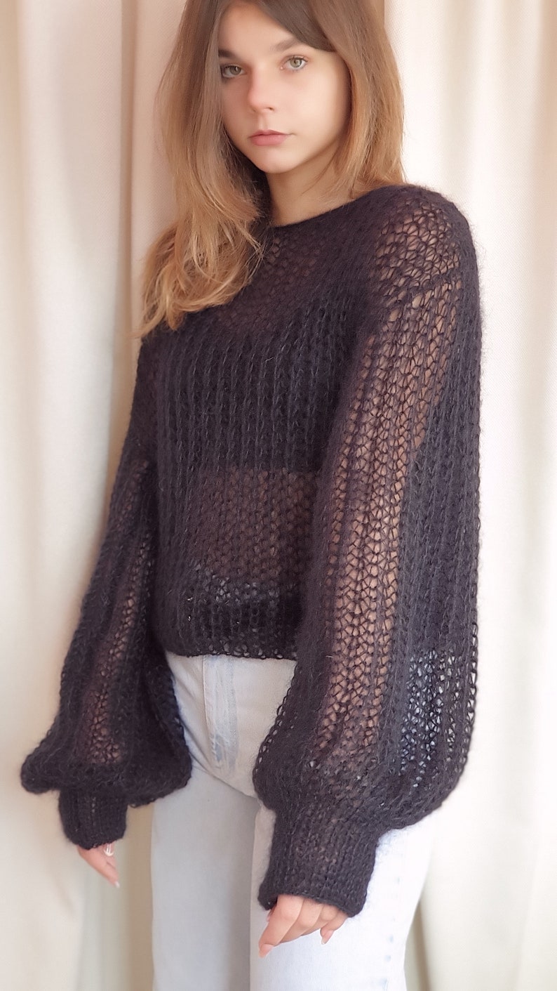 May include: A black, sheer, knitted sweater with long, puffed sleeves. The sweater is loose-fitting and cropped.