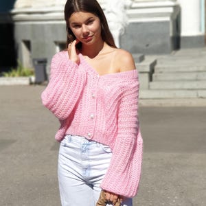 May include: A pink knitted cardigan with pearl buttons and long sleeves. The cardigan is cropped and has a slightly off-the-shoulder style. The model is wearing white jeans and a brown chain bag.