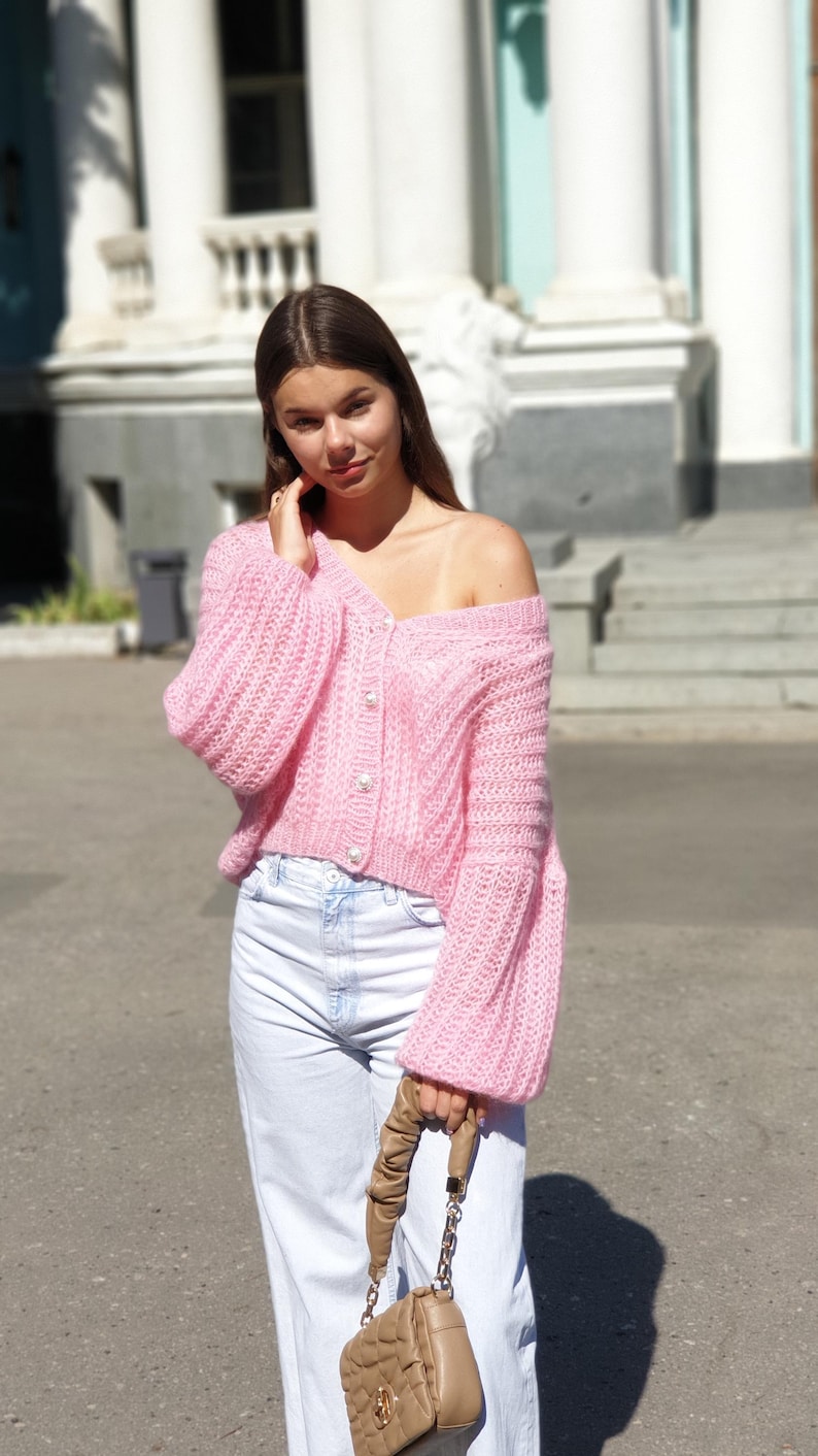 May include: A pink knitted cardigan with pearl buttons and long sleeves. The cardigan is cropped and has a slightly off-the-shoulder style. The model is wearing white jeans and a brown chain bag.