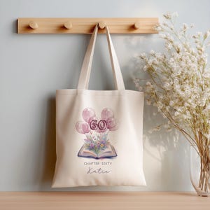 May include: A cream-coloured canvas tote bag featuring a watercolour illustration of an open book with a floral bouquet, topped by pink balloons forming the number 60. The text reads CHAPTER SIXTY and the name Katie in a script font below the book.