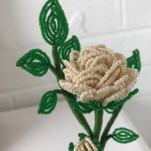 May include: A white beaded rose with green beaded leaves. The rose is made of small white beads and the leaves are made of small green beads. The rose is on a green stem.