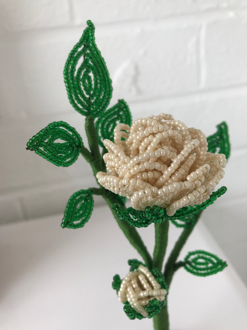 May include: A white beaded rose with green beaded leaves. The rose is made of small white beads and the leaves are made of small green beads. The rose is on a green stem.